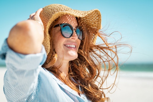 Redheaded woman on the beach with a hat and sunglasses on after eyelid surgery